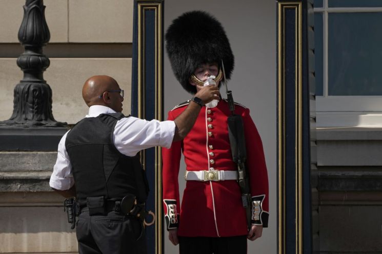 18일(현지 시각) 폭염이 닥친 영국 런던의 버킹엄궁 밖에서 한 경찰관이 곰털 모자를 쓰고 근무하는 왕실 근위병에게 물을 주고 있다. [이미지출처=연합뉴스]