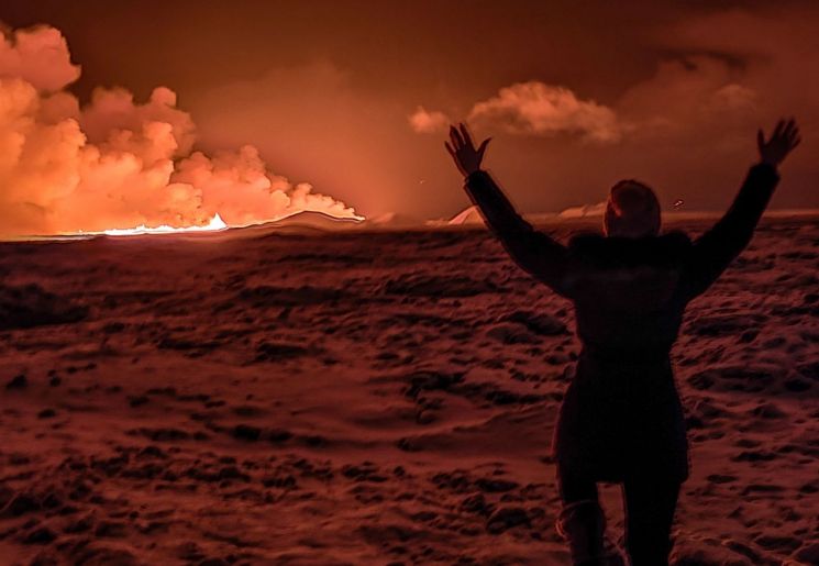 지난 18일(현지시간) 아이슬란드 남서부 그린다비크 인근 레이캬네스 반도에서 화산이 폭발해 밤하늘이 오렌지색으로 물든 모습을 한 주민이 두 팔을 치켜들고 바라보고 있다. [이미지출처=AFP연합뉴스]