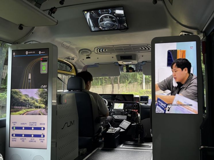 Inside Dongjak A01. The operator (safety officer) is seated in the driver's seat, and a camera screen showing the steering wheel is located at the top of the bus. The vertically oriented screen on the left displays the front camera, driving route, and surrounding objects, while the screen on the right plays news from Dongjak District Office and public information content. Photo by Jeon Youngju