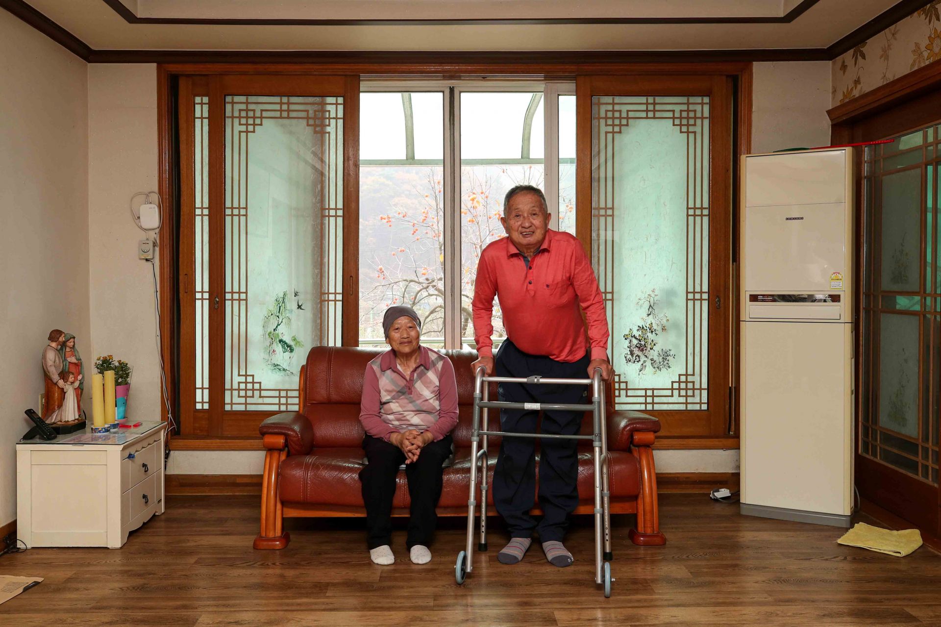 정학영(78) 할아버지와 김수남(76) 할머니가 충북 진천군 자택에서 카메라를 향해 포즈를 취하고 있다. 몸이 불편한 정학영 할아버지와 혈액암을 앓고 있는 김수남 할머니는 노후를 집에서 보내고 싶어 한다. 정학영, 김수남 부부의 진천 장군산 자락에 자리한 2층 집은 정 할아버지가 본인의 토지에 직접 건축한 집이다.