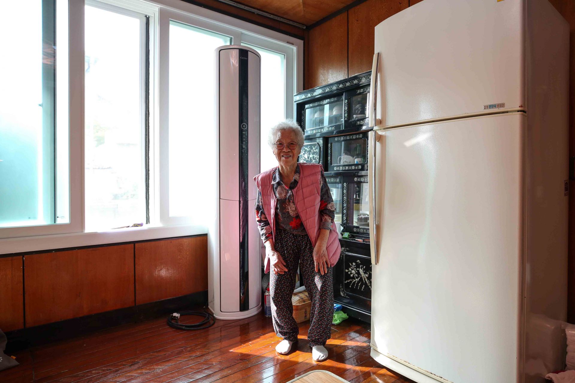 이정숙(92) 할머니가 충북 진천군 자택에서 아들이 설치해준 거실 에어컨 옆에 서서 카메라를 향해 포즈를 취하고 있다. 할머니는 읍내 병원에서 갈비뼈 수술을 받고 군청이 운영하는 퇴원 후 돌봄 서비스를 받으며 생활하고 있다.
